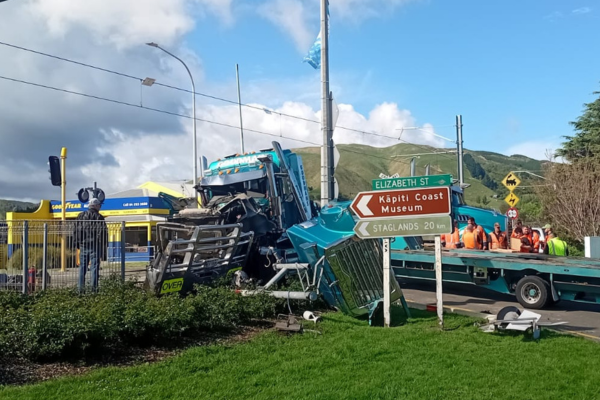Crash scene showing a damaged blue truck after a collision with a freight train near Waikanae Railway Station, with emergency workers on site and road signs pointing to Elizabeth Street and Kāpiti Coast Museum.